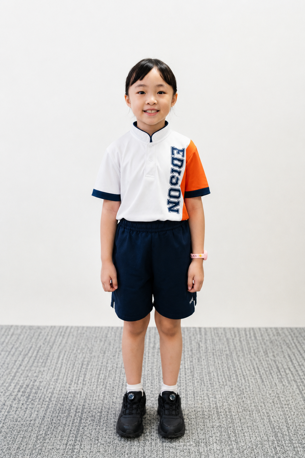 Smiling girl in white and orange school polo and navy shorts, standing on gray carpet against white wall.